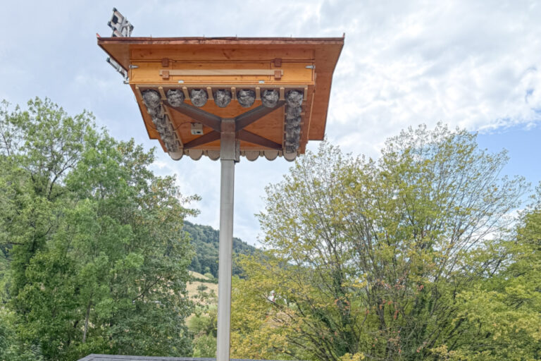 Fünf Personen in rot-gelber Kleidung stehen nebeneinander vor einem Pavillon unter einem hohen Holzmast mit Dach.