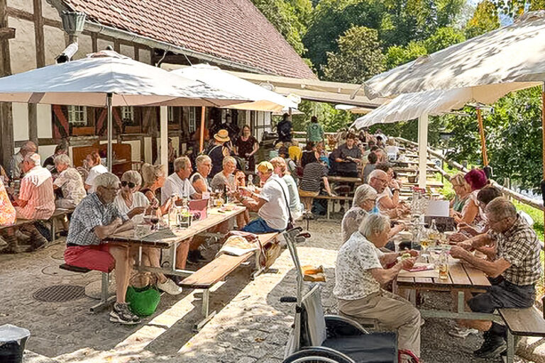 Menschen sitzen an Tischen unter Sonnenschirmen vor einem Fachwerkhaus im Freien und essen.