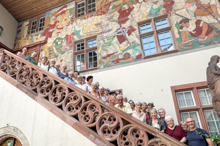 Eine grosse Gruppe steht lächelnd auf einer geschwungenen Freitreppe vor einem bemalten Fassadengiebel.