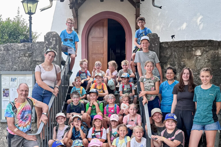 Grosse Gruppe von Kindern und Erwachsenen sitzt und steht auf einer Treppe vor einem Kirchenportal.