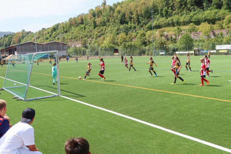 Junge Fussballerinnen kämpfen auf einem Kunstrasenplatz um den Ball, im Hintergrund bewaldete Hügel.