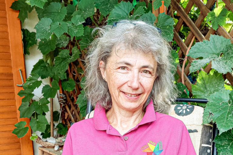 Eine Frau mit grauen Haaren sitzt lächelnd im Garten und hält ein türkises Heft mit der Aufschrift „Ferienpass“.
