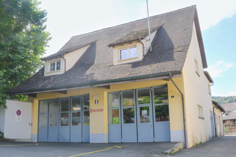 Ein gelbes Gebäude mit grauen Falttoren und steilem Ziegeldach, davor ein leerer Parkplatz.