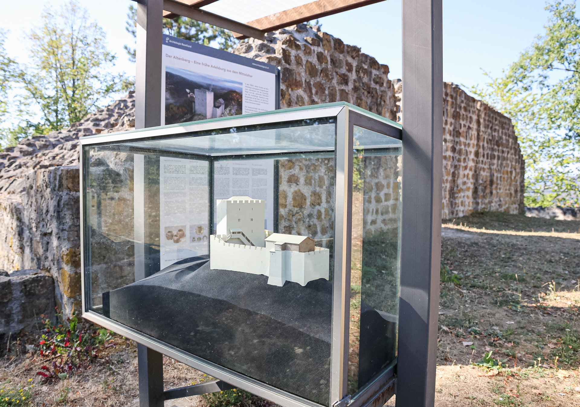 Ein Modell einer Ruine steht in einer Glasvitrine vor einer historischen Steinmauer im Freien.