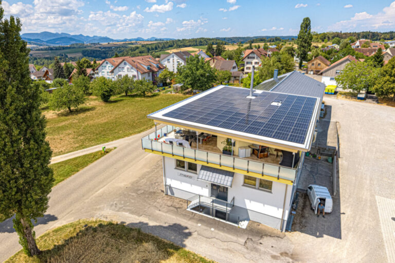 Luftaufnahme eines Gebäudes mit grossflächiger Photovoltaikanlage auf dem Dach, umgeben von grüner Landschaft.
