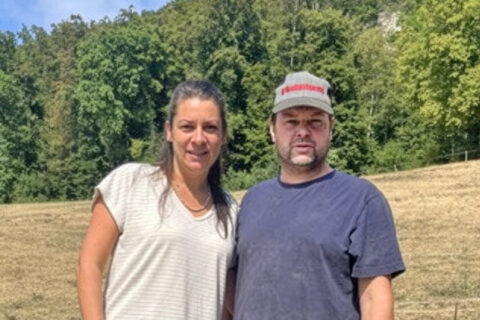 Eine Frau und ein Mann stehen nebeneinander auf einer trockenen Wiese vor bewaldeten Hügeln.
