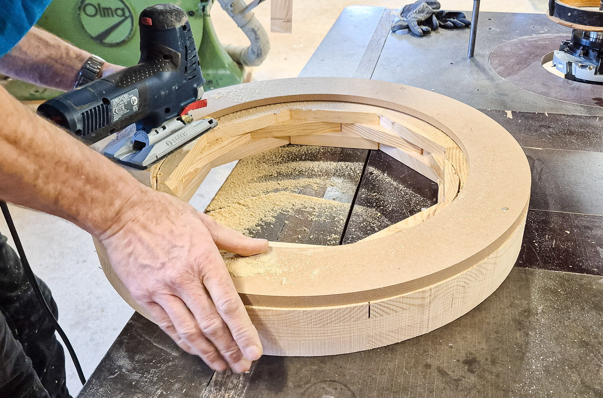 Eine Hand hält einen runden Holzring auf einer Werkbank fest, daneben Sägestaub und Werkzeug.