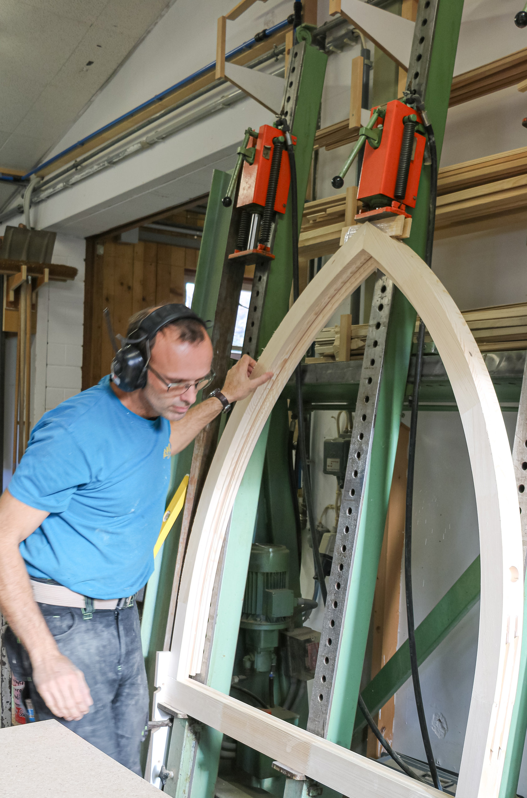 Ein Mann mit Gehörschutz betrachtet in einer Werkstatt einen gebogenen Holzrahmen in einer Spannvorrichtung.