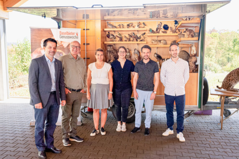 Sechs Personen stehen lächelnd nebeneinander vor einem Anhänger mit ausgestellten präparierten Tieren.