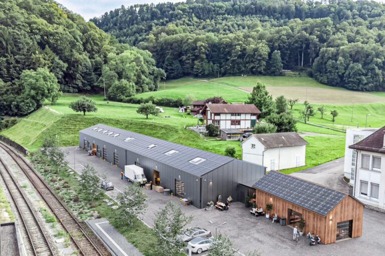 Luftaufnahme einer langen Lagerhalle mit Solaranlage auf dem Dach, umgeben von Wiesen und Wald.