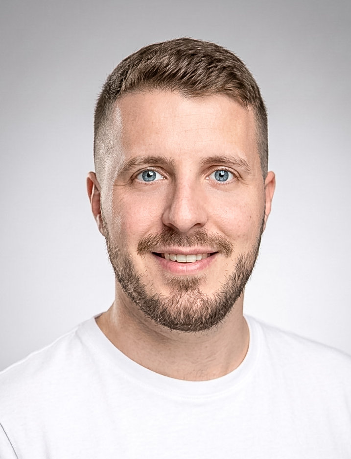 Ein lächelnder junger Mann mit kurzen braunen Haaren und Bart blickt in die Kamera.