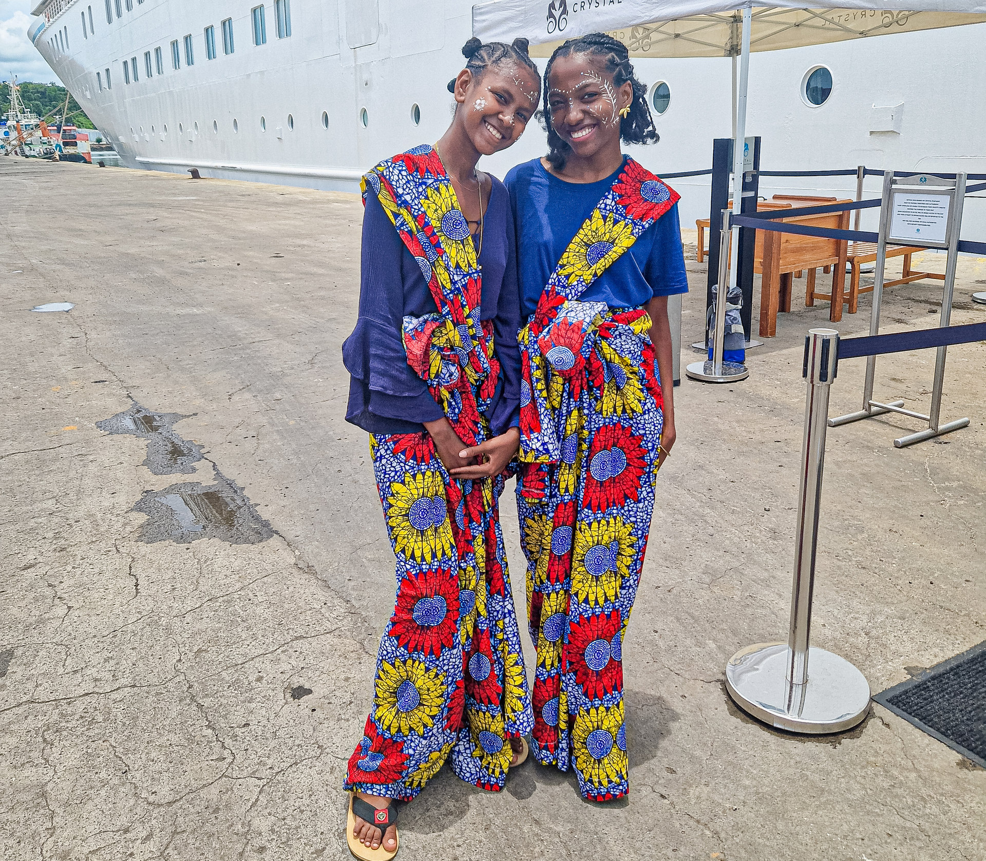 Zwei lächelnde junge Frauen in bunten gemusterten Gewändern stehen eng nebeneinander vor einem Kreuzfahrtschiff.