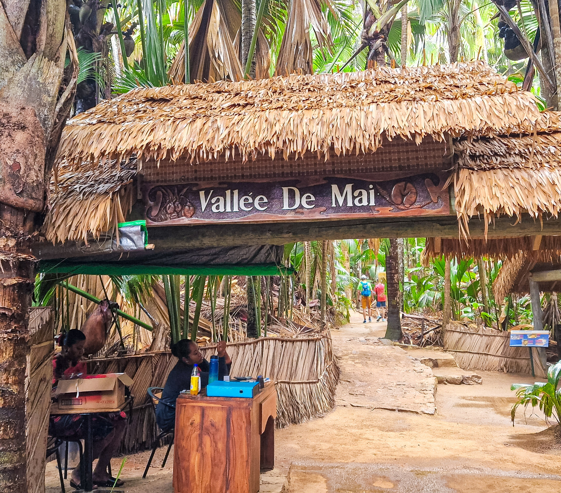 Ein strohgedecktes Torgebäude mit der Aufschrift „Vallée De Mai“, umgeben von tropischem Palmenwald.