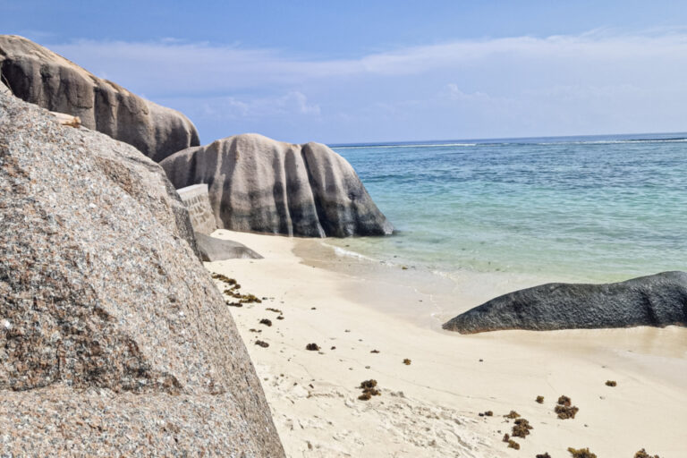 Grosse, glatt geformte Granitfelsen ragen an einem weissen Sandstrand neben türkisfarbenem Meer empor.