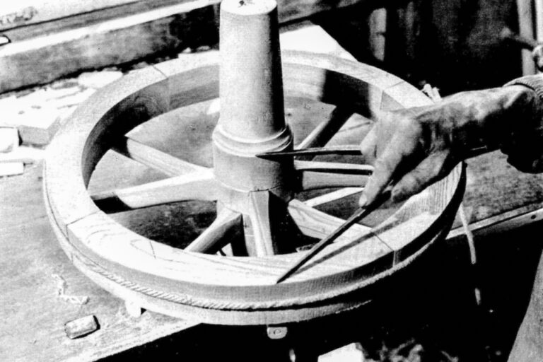 Eine Hand hält einen Zirkel an ein hölzernes Wagenrad mit Speichen und zentraler Nabe.