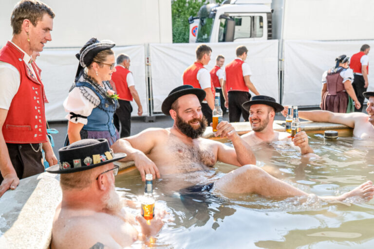 Männer mit Trachtenhüten sitzen lachend und bierselig in einem Wasserbecken, im Hintergrund Trachtenträger.