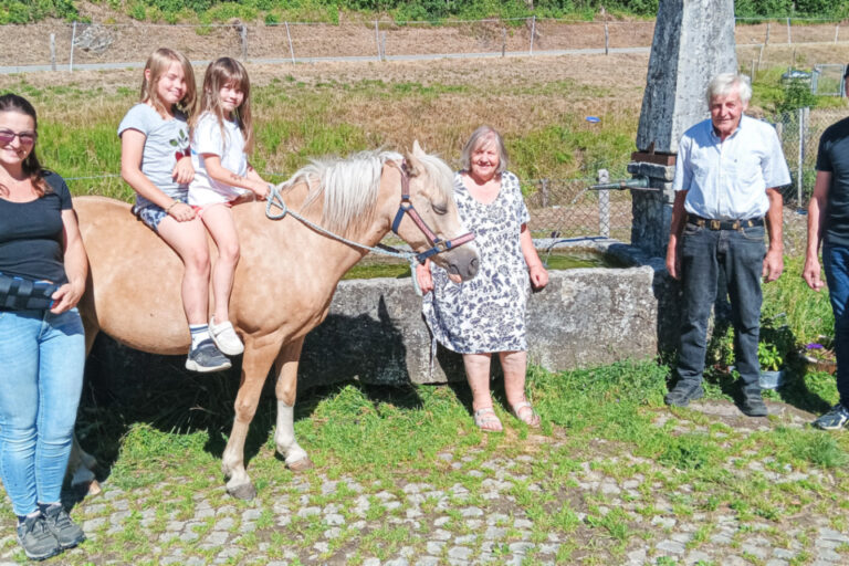 Sechs Personen stehen bei einem Steintrog, zwei Mädchen sitzen auf einem Pony, im Hintergrund Grün und ein steinernes Denkmal.