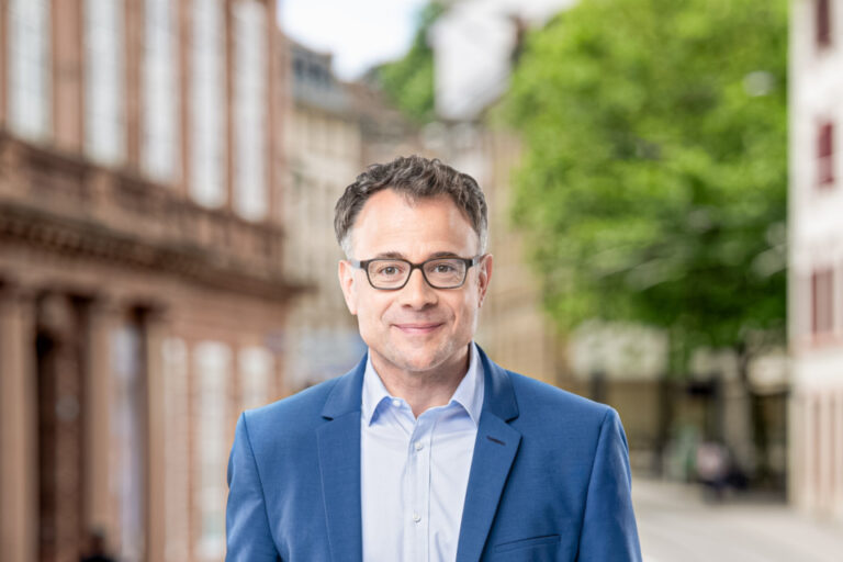 Ein lächelnder Mann mit Brille und blauem Sakko steht auf einer Straße in der Altstadt.