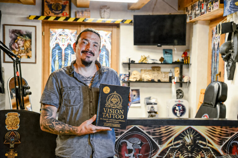 Ein tätowierter Mann mit Bart hält lächelnd ein Buch mit dem Titel «Vision Tattoo» in einem Tattoostudio.