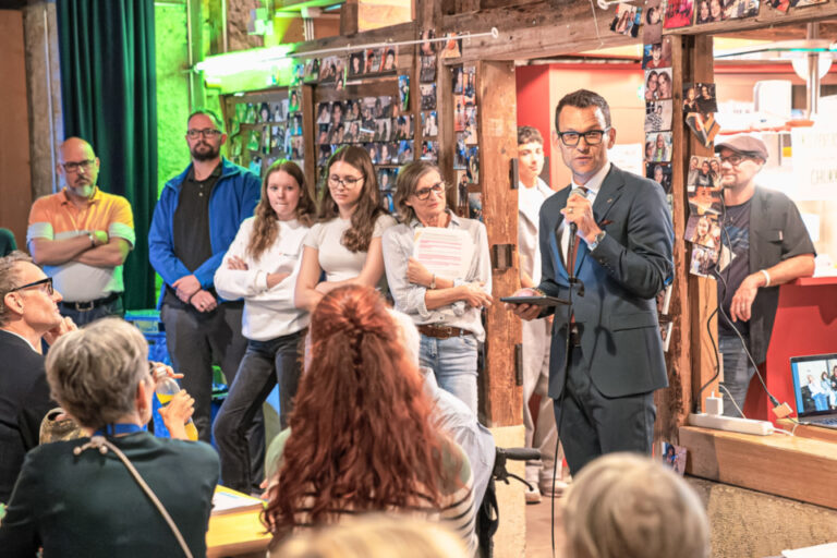 Ein Mann im Anzug spricht mit Mikrofon vor stehenden Zuhörern in einem mit Fotos dekorierten Raum.