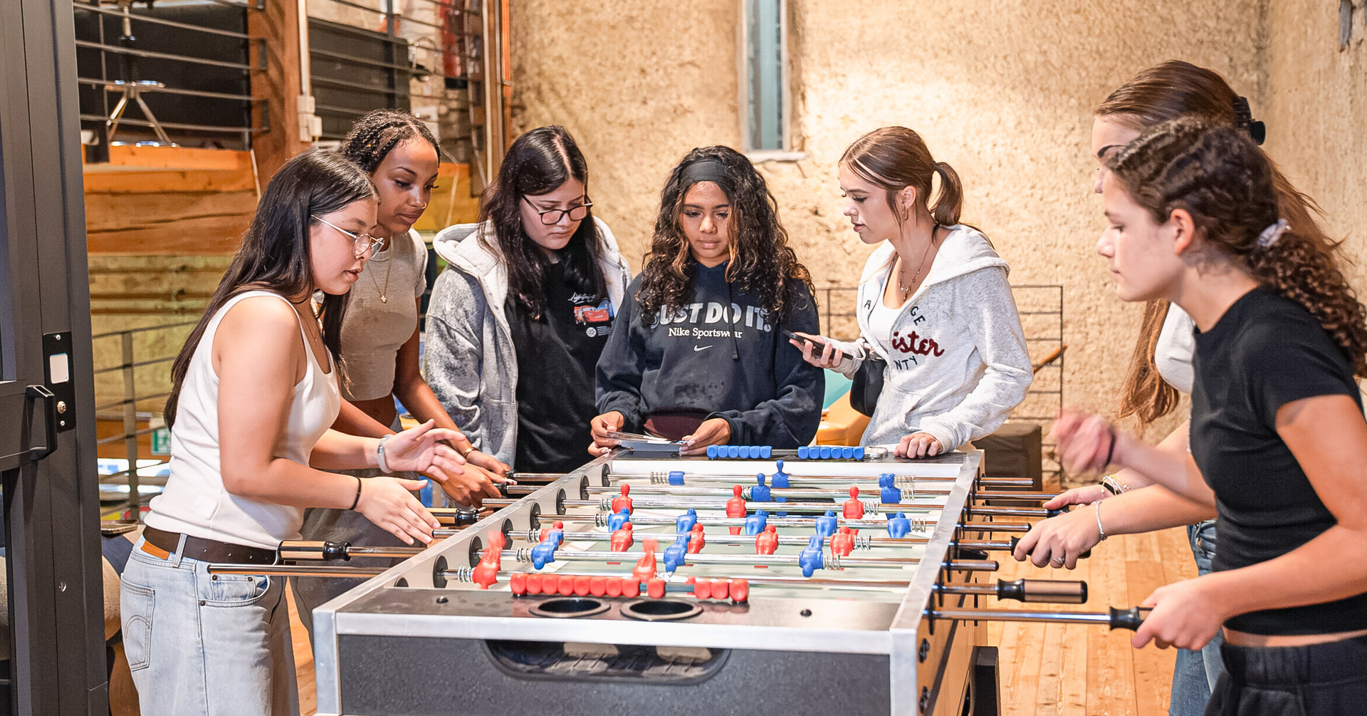 Sechs junge Frauen stehen lachend um einen Tischfussballtisch in einem rustikalen Raum.