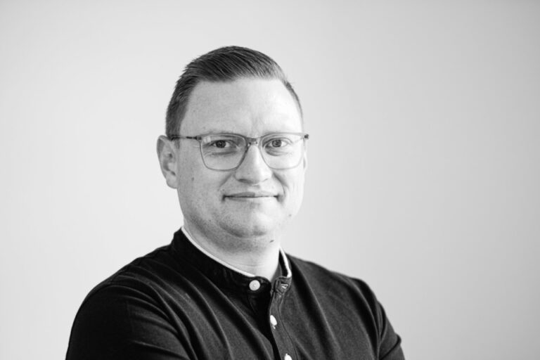 Schwarz-weiss Porträt eines lächelnden Mannes mit Brille und dunklem Hemd vor hellem Hintergrund.