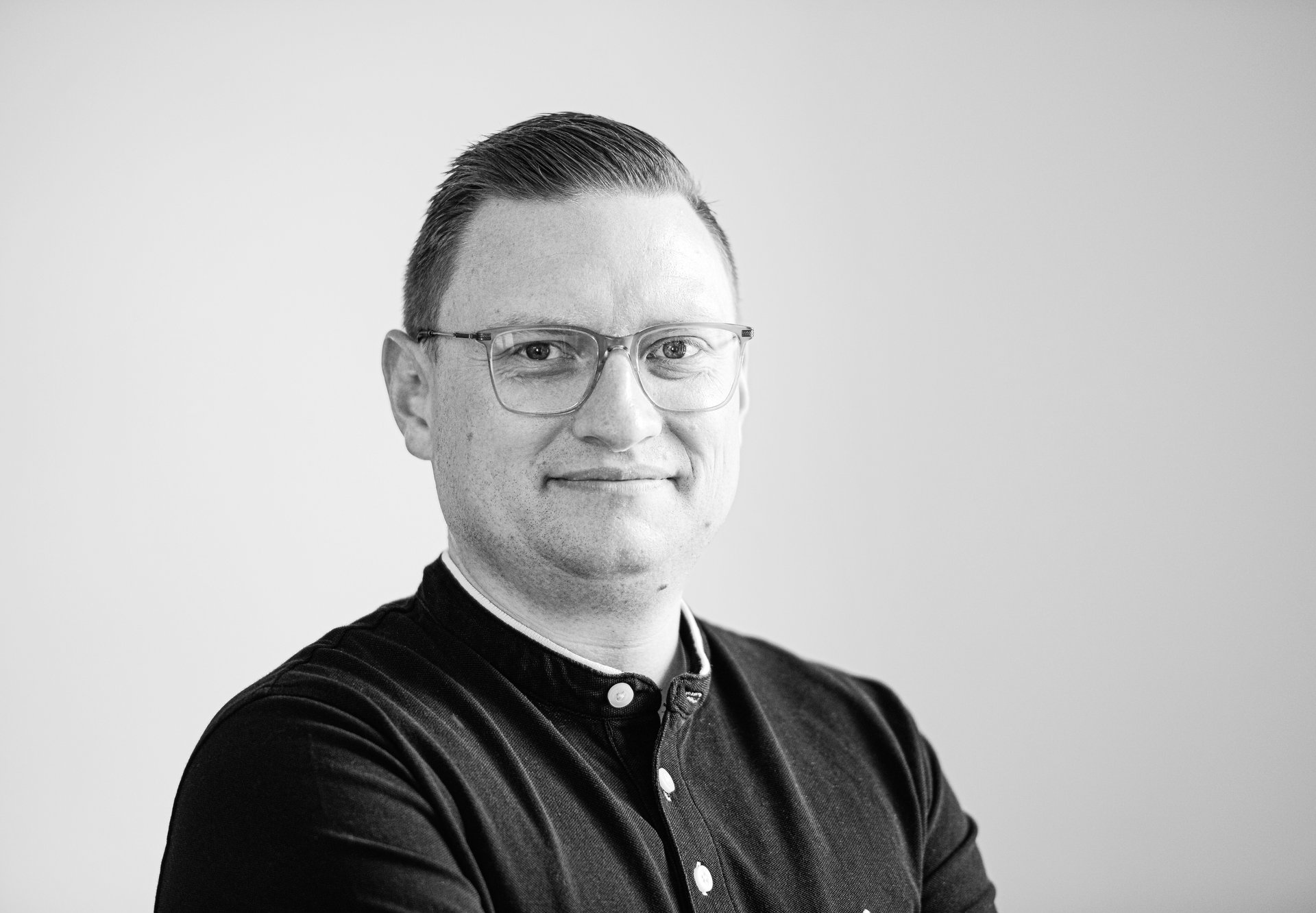 Schwarz-weiss Porträt eines lächelnden Mannes mit Brille und dunklem Hemd vor hellem Hintergrund.