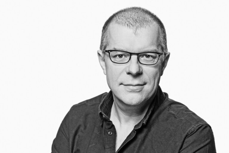 Schwarz-weiß-Porträt eines kurzhaarigen Mannes mit Brille und dunklem Hemd, der in die Kamera blickt.