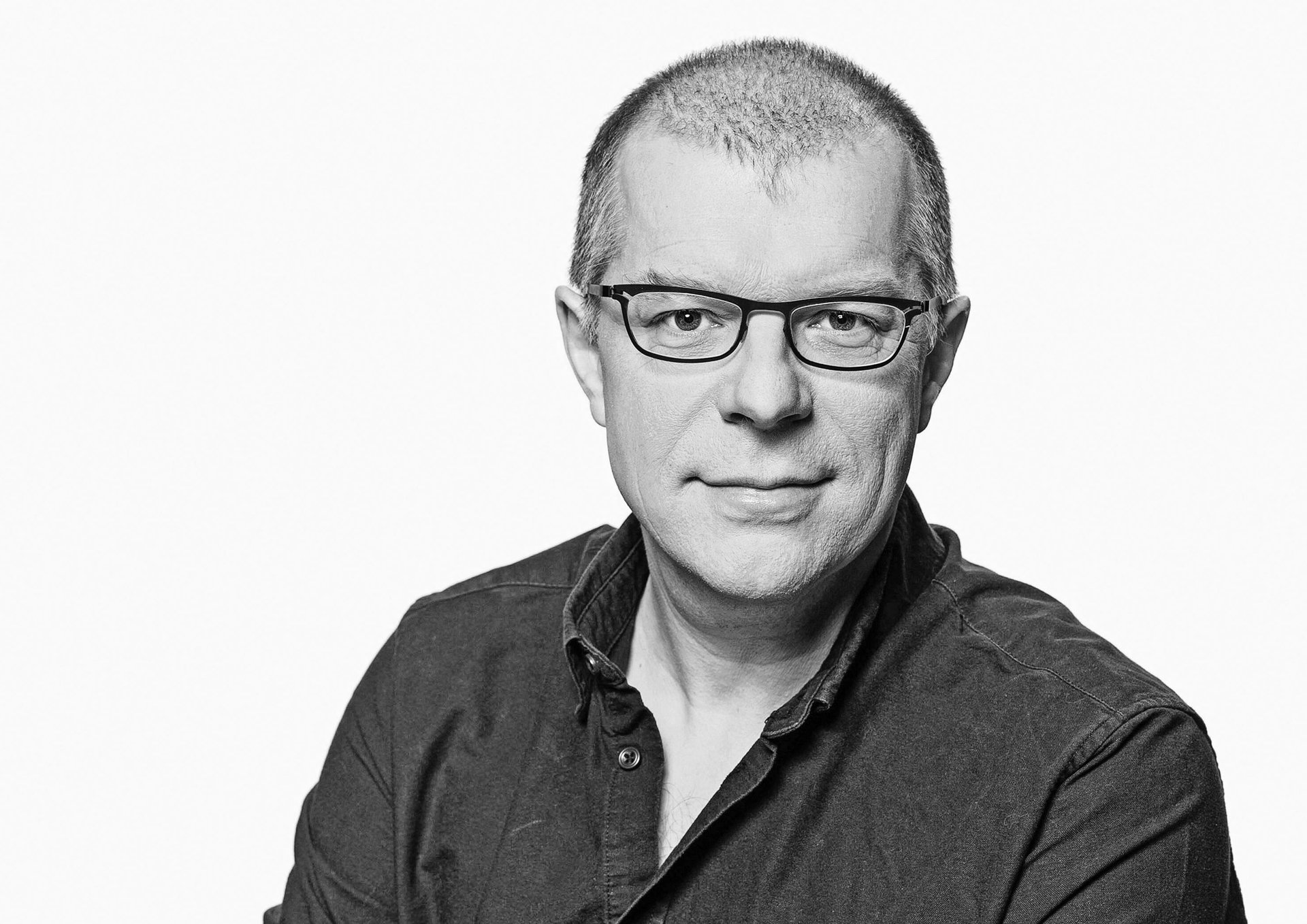 Schwarz-weiß-Porträt eines kurzhaarigen Mannes mit Brille und dunklem Hemd, der in die Kamera blickt.