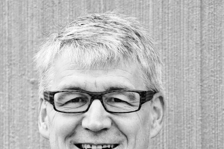 Ein älterer Mann mit grauem Haar und Brille lächelt, er trägt ein dunkles Hemd.
