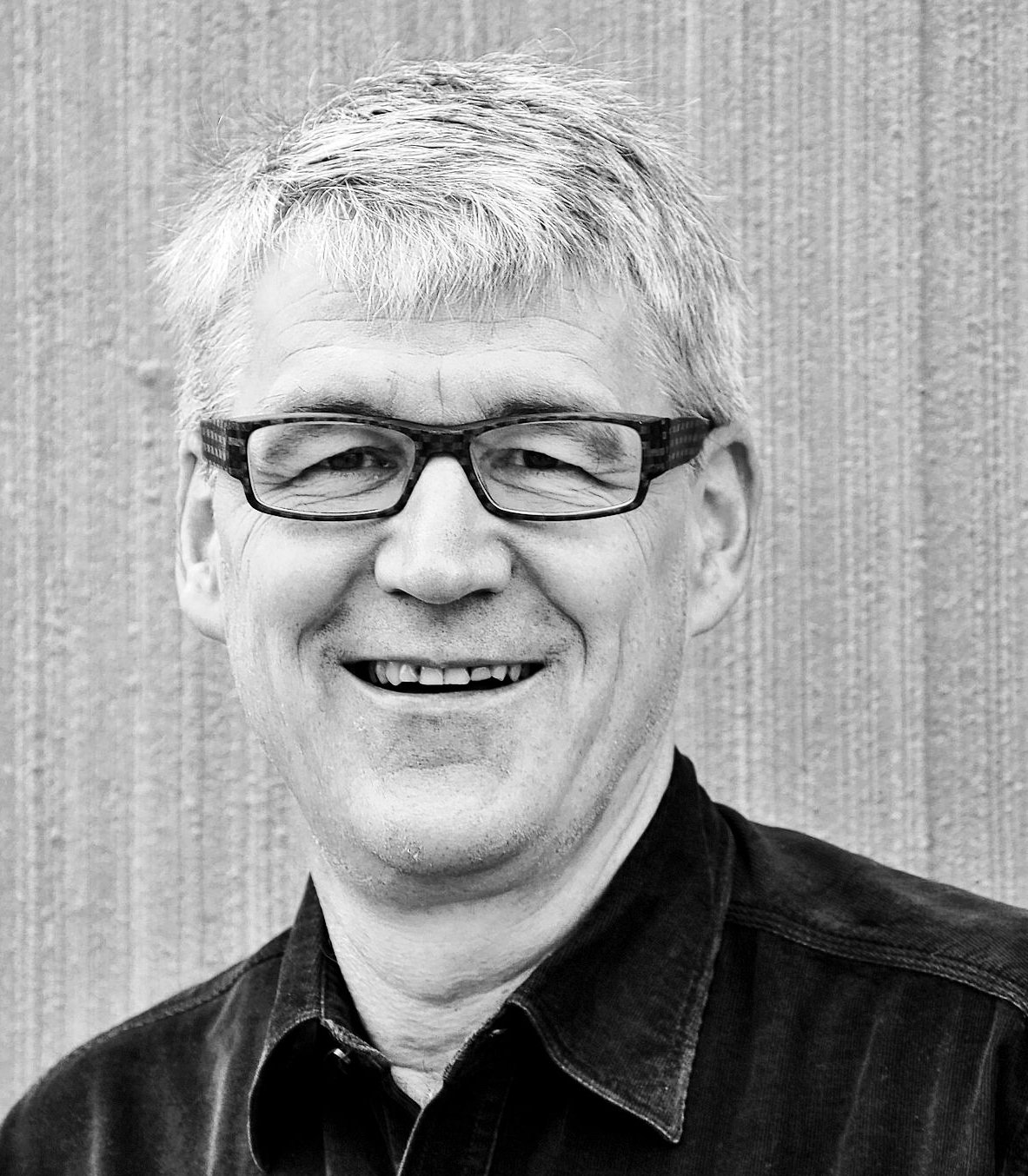 Ein älterer Mann mit grauem Haar und Brille lächelt, er trägt ein dunkles Hemd.