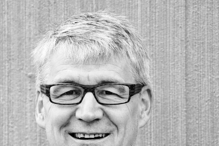 Ein lächelnder Mann mittleren Alters mit Brille und grauem Haar in dunklem Hemd vor gestreifter Wand.