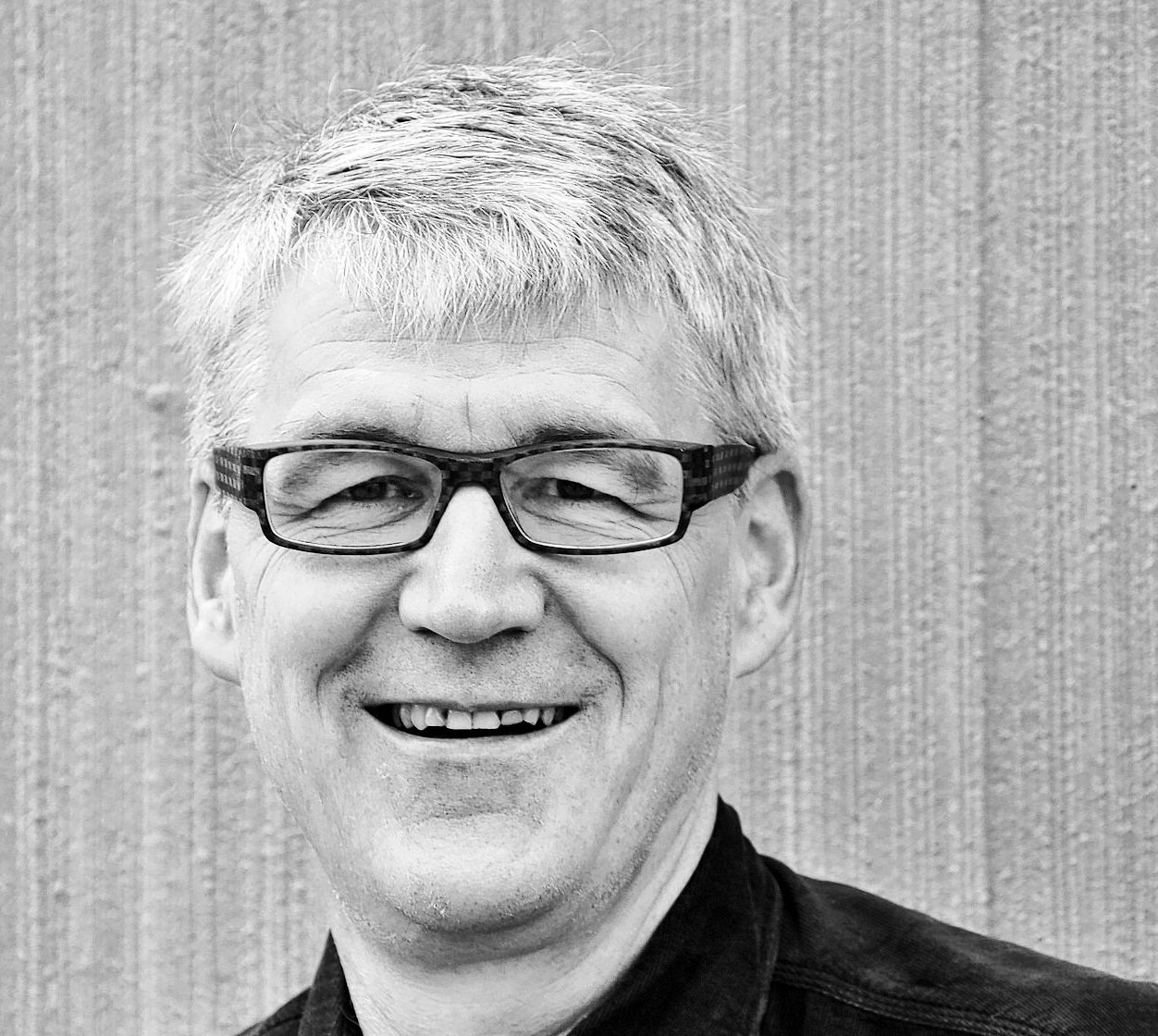 Ein lächelnder Mann mittleren Alters mit Brille und grauem Haar in dunklem Hemd vor gestreifter Wand.