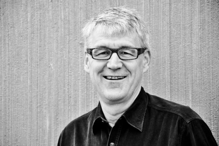 Ein lächelnder älterer Mann mit Brille und grauem Haar in dunklem Hemd vor gestreifter Wand.