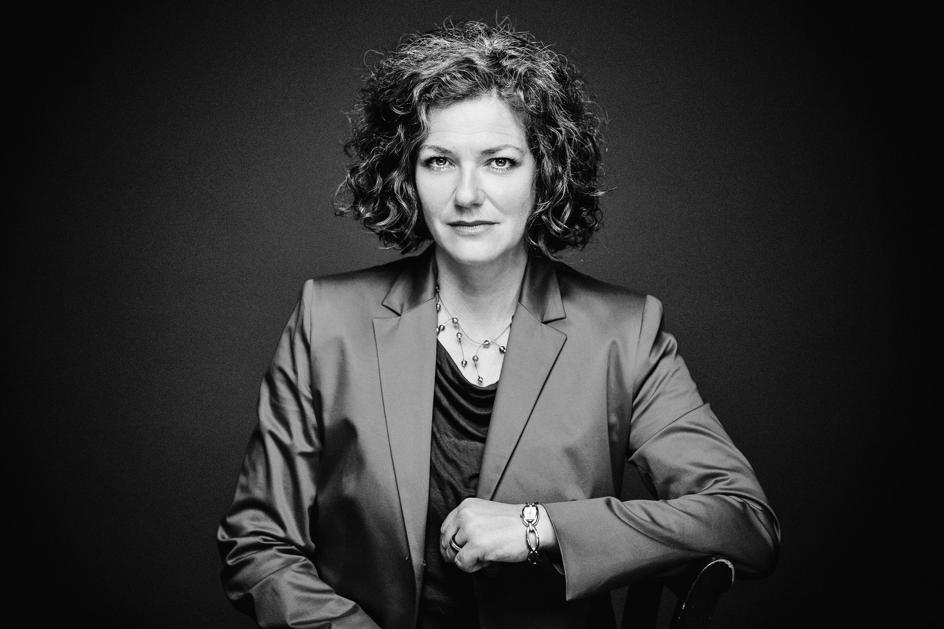 Schwarz-weiß-Porträt einer Frau mit lockigem Haar und Blazer, die in die Kamera blickt.
