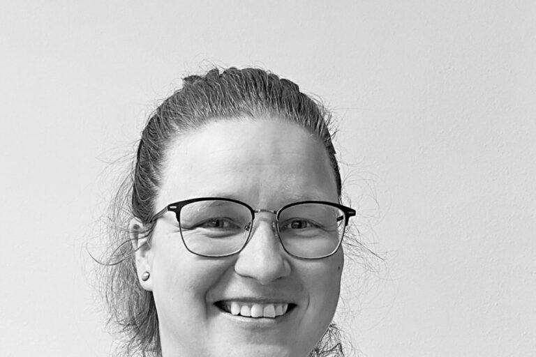 Eine lächelnde Frau mittleren Alters mit Brille und zurückgebundenem Haar in Schwarz-Weiß.
