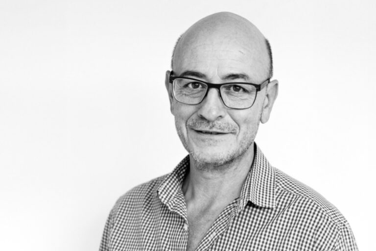 Ein älterer Mann mit Brille, Halbglatze und kariertem Hemd blickt lächelnd in die Kamera.