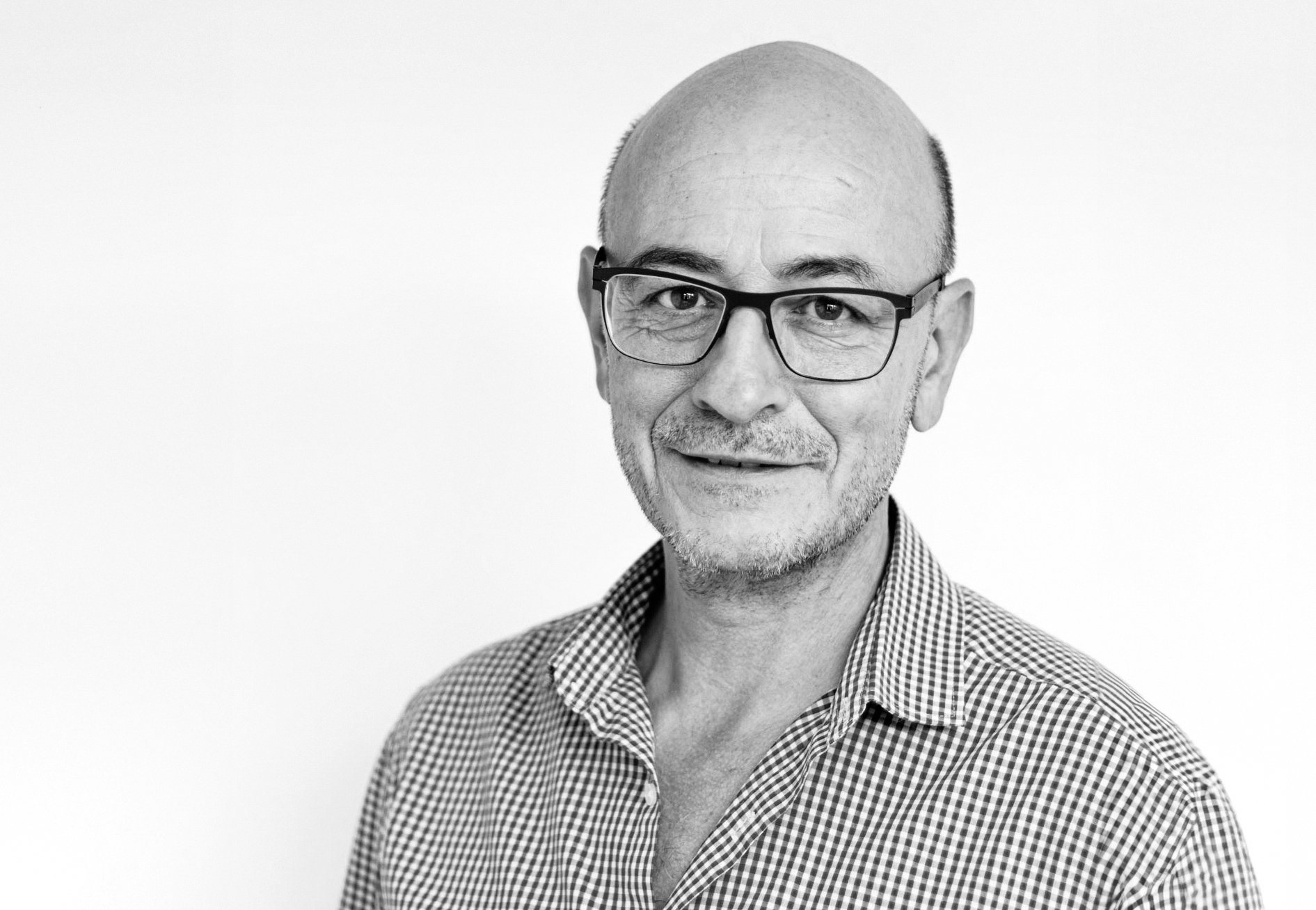 Ein älterer Mann mit Brille, Halbglatze und kariertem Hemd blickt lächelnd in die Kamera.