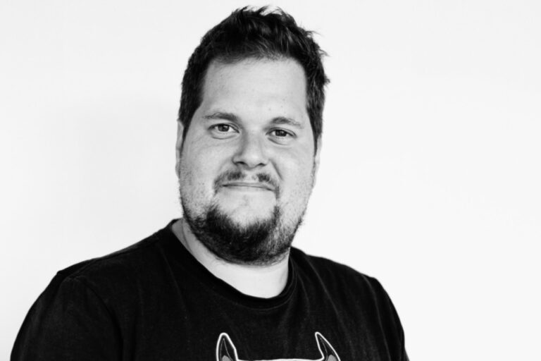 Schwarz-weiss-Porträt eines lächelnden Mannes mit Bart und dunklem T-Shirt vor heller Wand.
