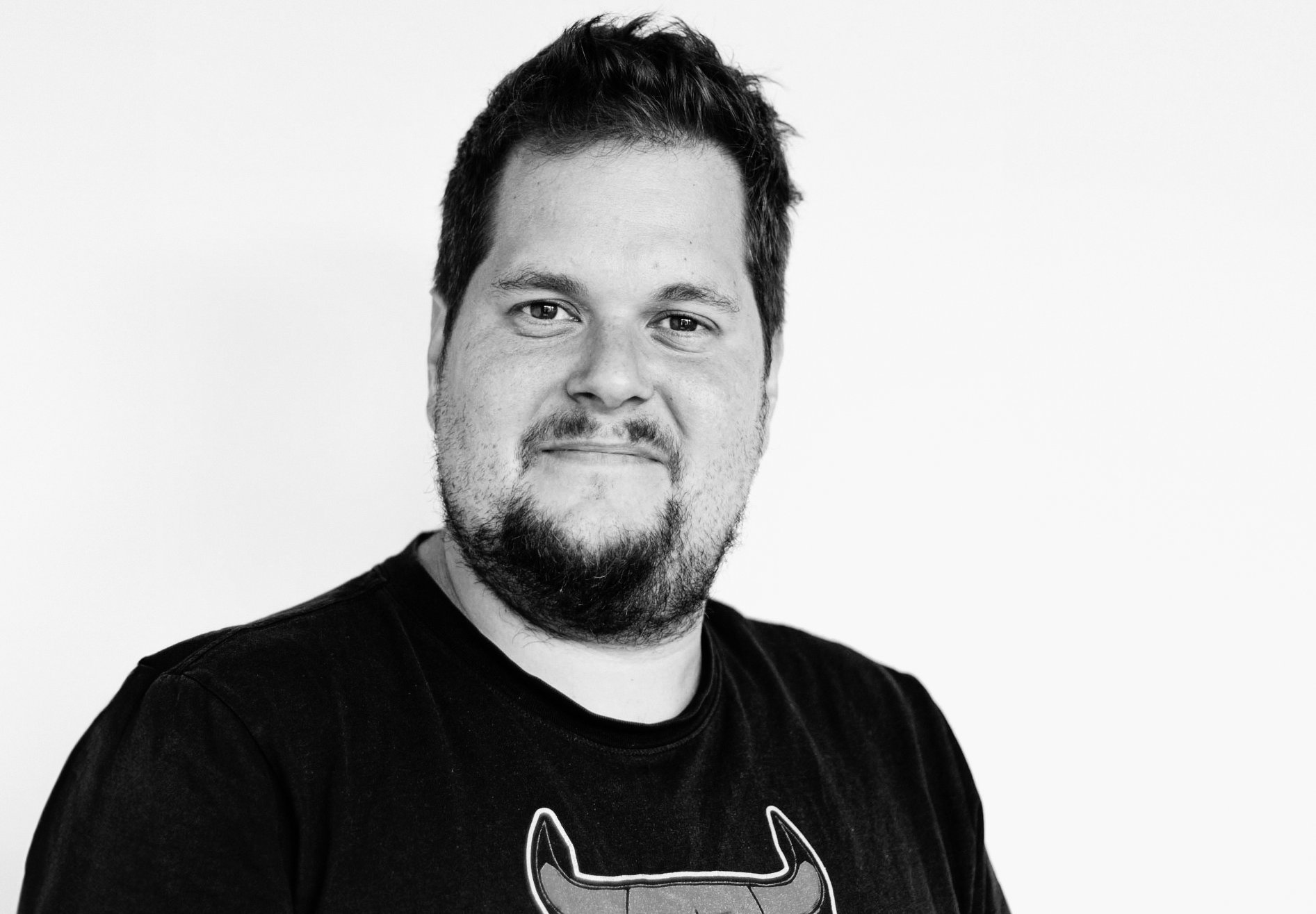 Schwarz-weiss-Porträt eines lächelnden Mannes mit Bart und dunklem T-Shirt vor heller Wand.