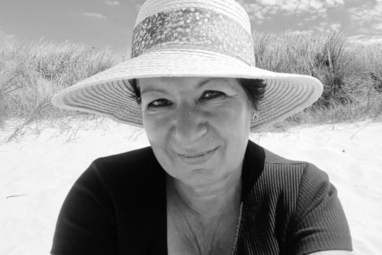 Lächelnde Frau mit Sonnenhut sitzt am Strand vor Dünengras, Schwarzweißfoto.
