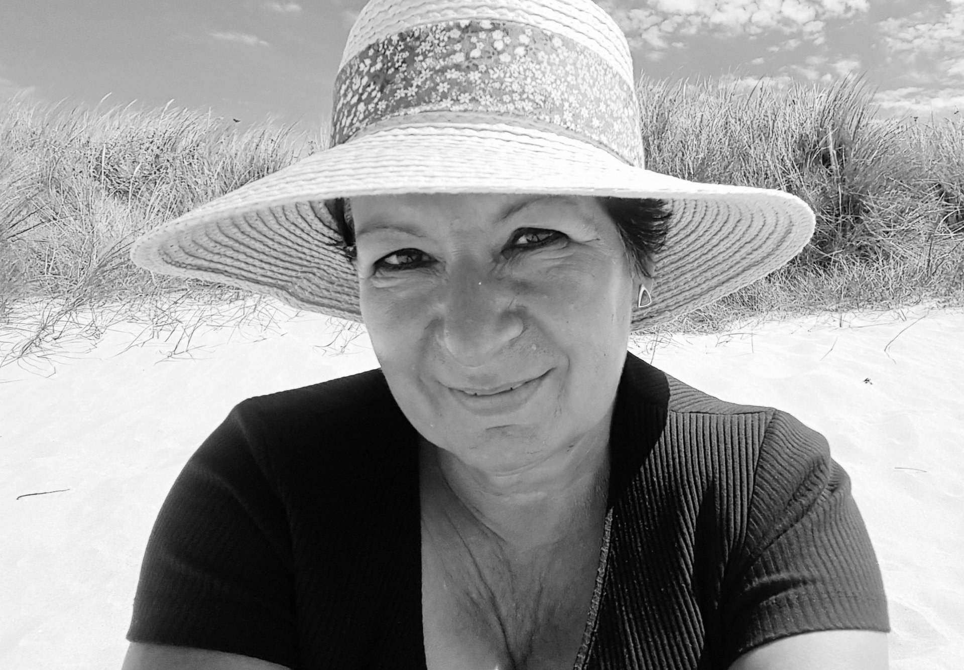 Lächelnde Frau mit Sonnenhut sitzt am Strand vor Dünengras, Schwarzweißfoto.