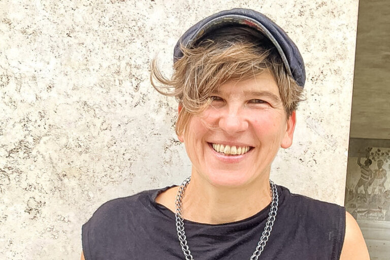 Lächelnde Frau mit kurzen Haaren und Schirmmütze, sie trägt ein dunkles Top und eine Kette.