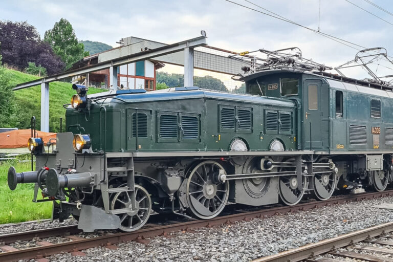 Eine dunkelgrüne historische Elektrolokomotive mit markanten Rädern steht auf Gleisen unter Fahrleitungen.