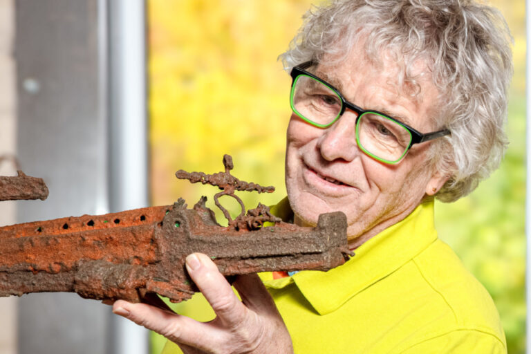 Ein lächelnder älterer Mann mit grüner Brille hält ein stark verrostetes altes Metallobjekt in den Händen.
