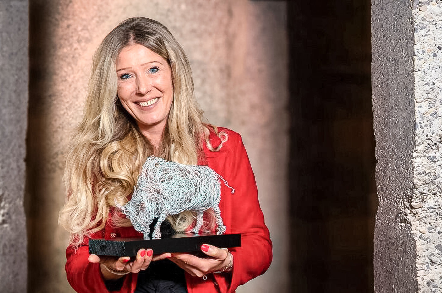 Lächelnde blonde Frau in rotem Blazer hält eine Skulptur aus Drahtgeflecht in Tierform.