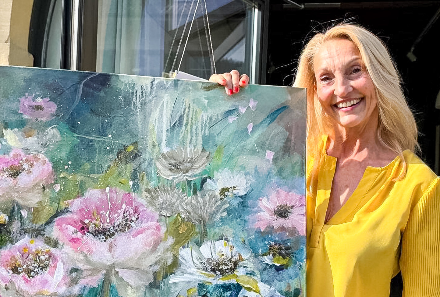 Eine lächelnde Frau mittleren Alters in gelber Bluse steht neben einem gemalten Blumenbild.