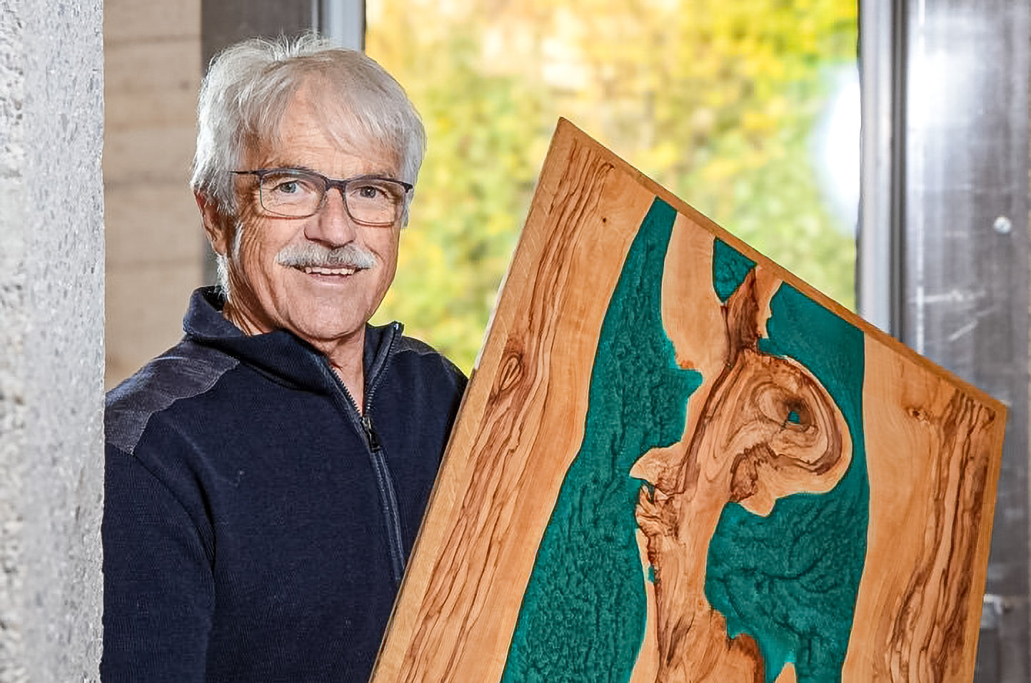 Ein älterer Mann mit grauem Haar und Brille lächelt und hält eine Holzplatte mit türkiser Kunstharz-Einlage.