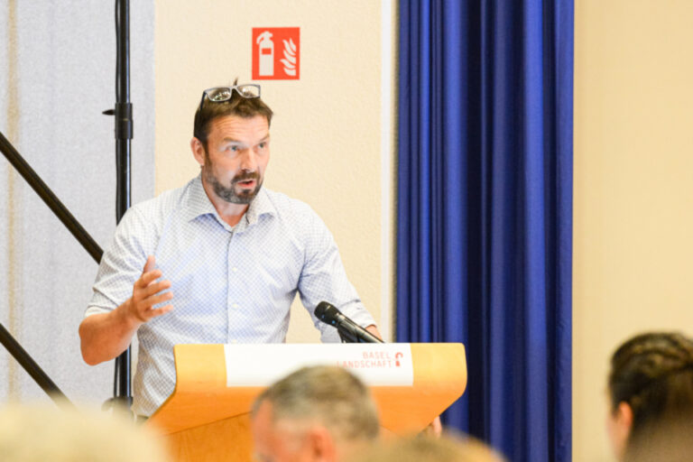 Ein Mann in hellem Hemd spricht gestikulierend an einem Rednerpult vor sitzendem Publikum in einem Saal.