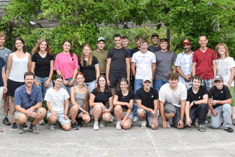 Gruppenfoto mit rund 25 jungen Menschen, stehend und kniend, vor grünen Bäumen auf einem Innenhof.